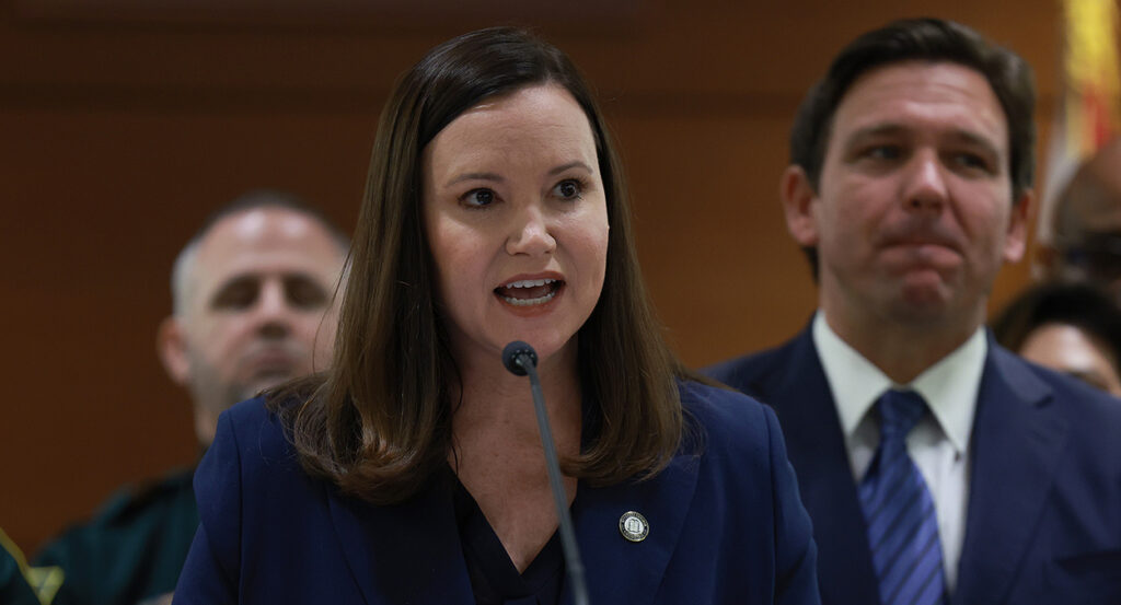 Ashley Moody in a blue suit speaks in front of Ron DeSantis in a blue suit.