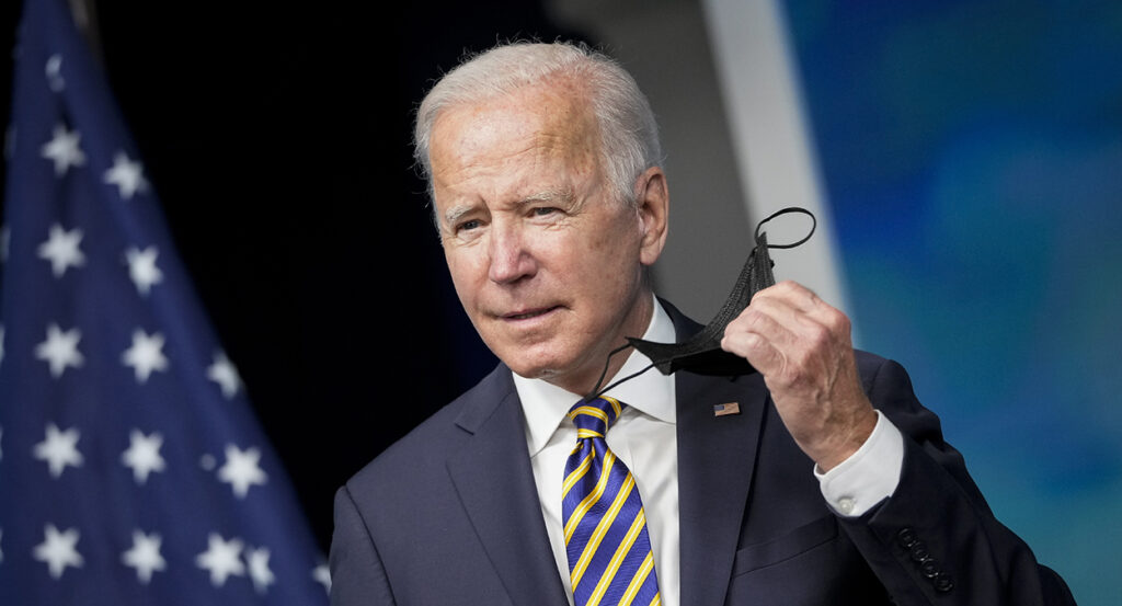 Joe Biden in a suit with an American flag pin holds a black mask in his left hand.