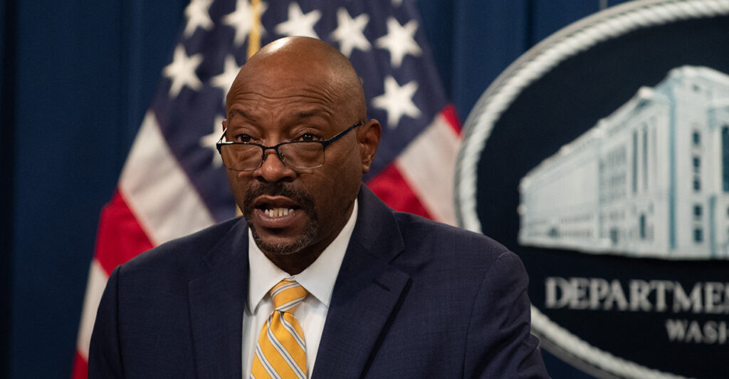 Tae Johnson speaks at a podium wearing a dark suit and yellow tie with the American flag behind him.