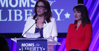 Tiffany Justice in a blue dress and white blazer speaks with Tina Descovich in a red dress in front of a Moms for Liberty logo