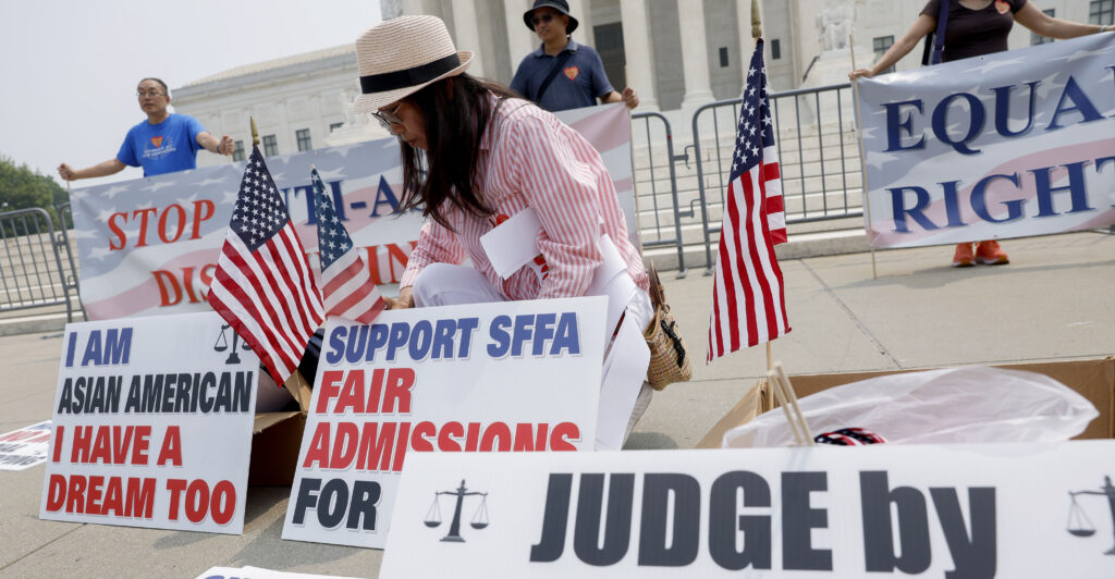 An Asian American woman wearing a red and white striped shirt and tan wicker hat kneels in front of the Supreme Court, surrounded by American flags and signs expressing anti-affirmative action sentiments. She is celebrating the day.