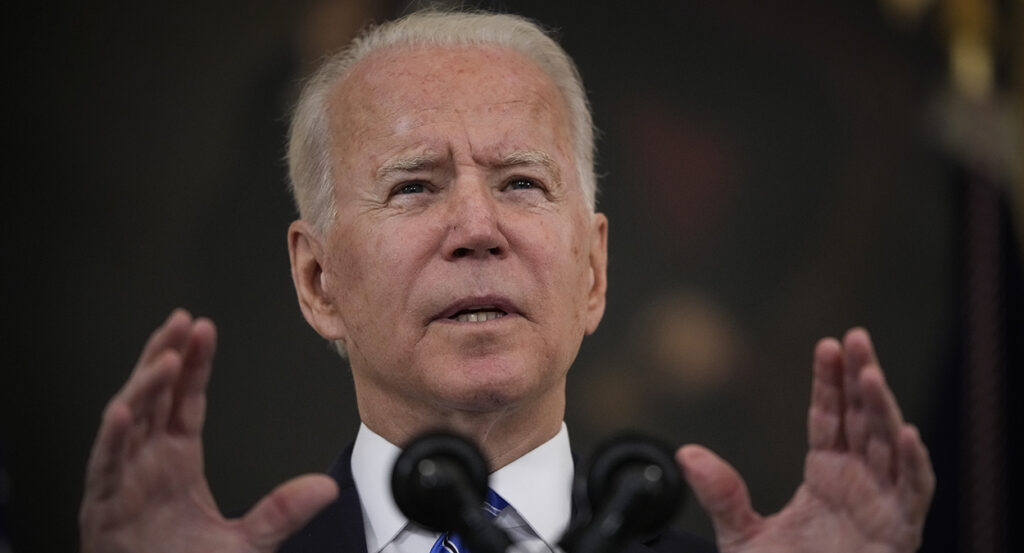 President Joe Biden gestures in front of a microphone while wearing a suit and a blue striped tie