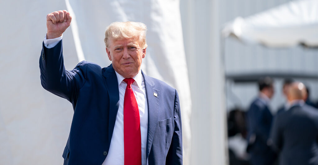 President Donald Trump greets a crowd raising his arm, wearing a black suit and a read tie