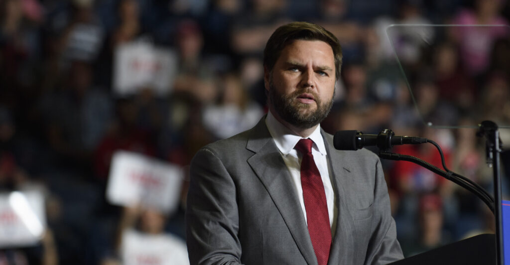 J.D. Vance in a gray suit with a red tie speaks in front of a crowd