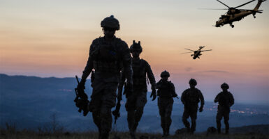 Five Army soldiers on the ground and two helicopters in the air at dusk