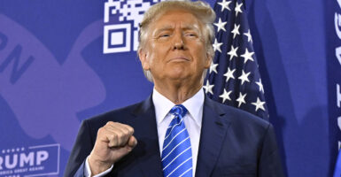 Donald Trump raises a fist with a grim smile on his face, wearing a blue suit in front of an American flag