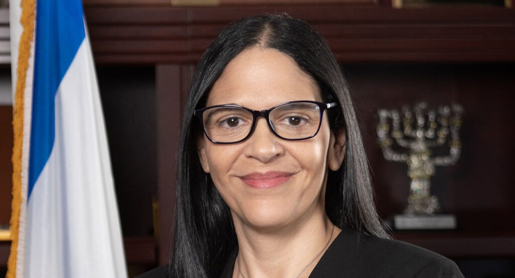 Anat Sultan-Dadon, consul general of Israel, smiles in front of a menorah and an Israeli flag.