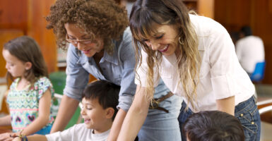 Two teachers, one wearing white and one wearing a blue jean shirt, help children learn