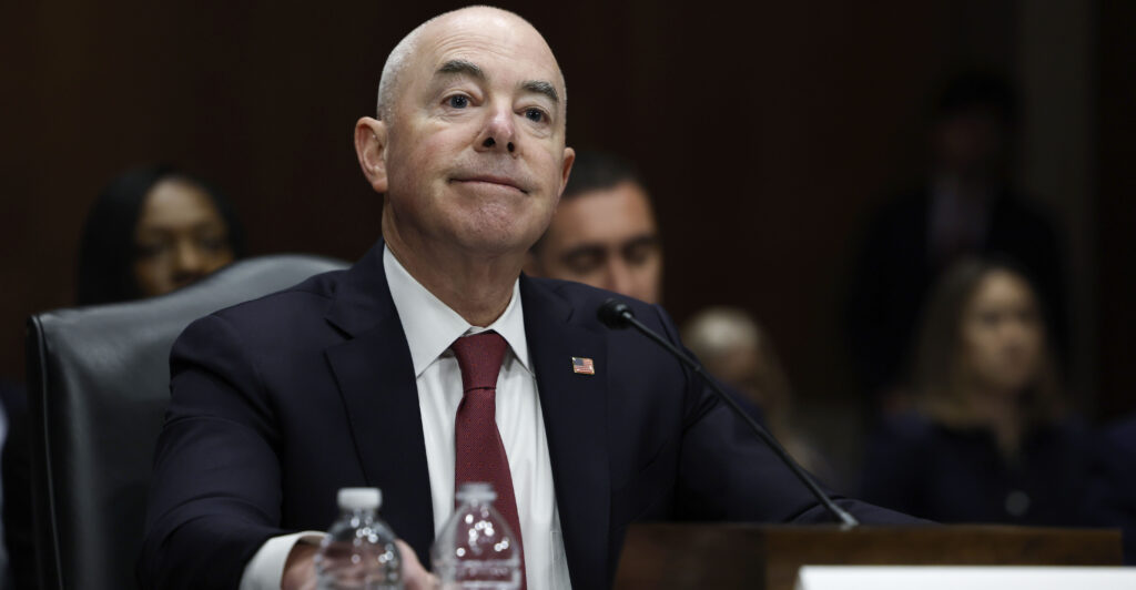 Mayorkas smiles while wearing a black suit and red tie.