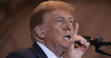 Former President Donald Trump in a suit at a microphone with his hand raised, Finger pointing upward