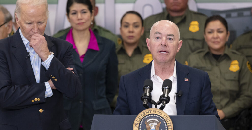 Alejandro Mayorkas stands behind a lectern in a navy blue suit, while President Joe Biden stands to the left of him.