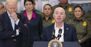 Alejandro Mayorkas stands behind a lectern in a navy blue suit, while President Joe Biden stands to the left of him.