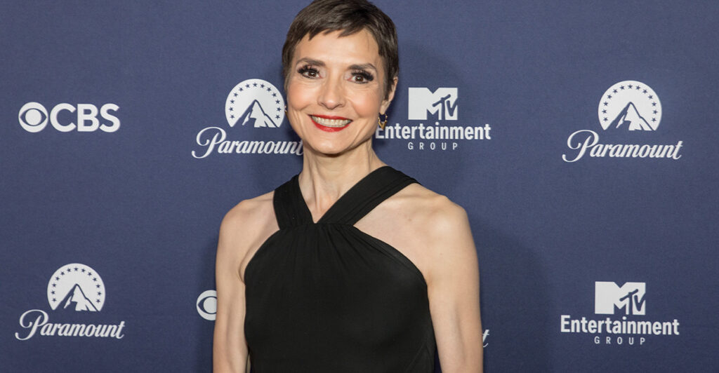 Catherine Herridge stand in front of a step and repeat in a black dress and smiles.