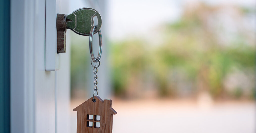 A key with a small wood house key chain hands from a white door.