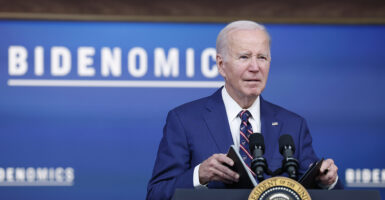 Joe Biden in a blue suit holds a briefing book behind the presidential seal and in front of a sign reading "Bidenomics."
