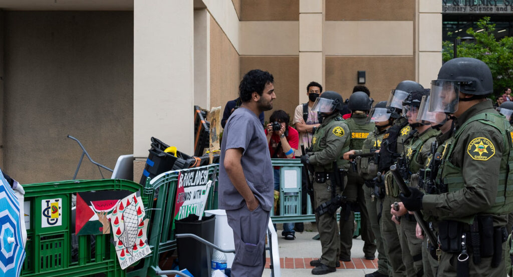 A protester wearing a gray shirt and jeans stands before a line of police officers wearing face shields and green uniforms.