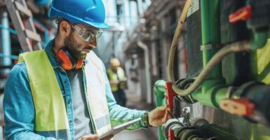 A young male engineer With a hard hat is examining the pipe system In a factory and checking the data on his tablet.