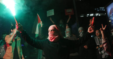 A man wearing a head and face covering holds a green flare in the air.