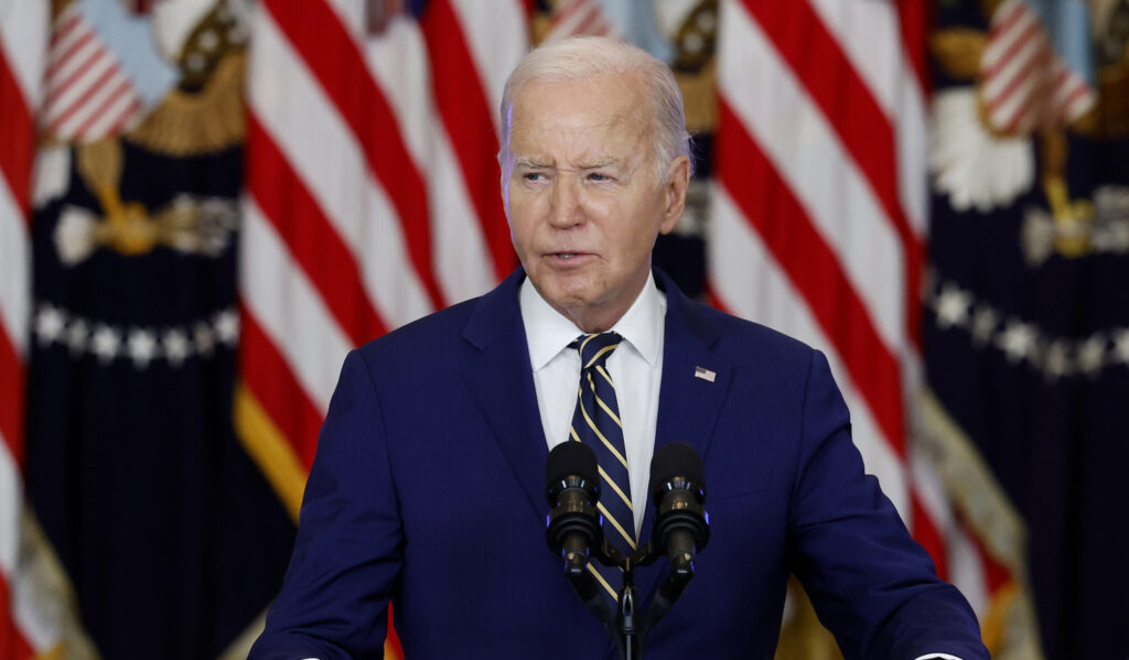 Joe Biden wears a navy-blue suit and speaks at a podium in front of American flags.