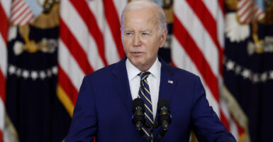 Joe Biden wears a navy-blue suit and speaks at a podium in front of American flags.