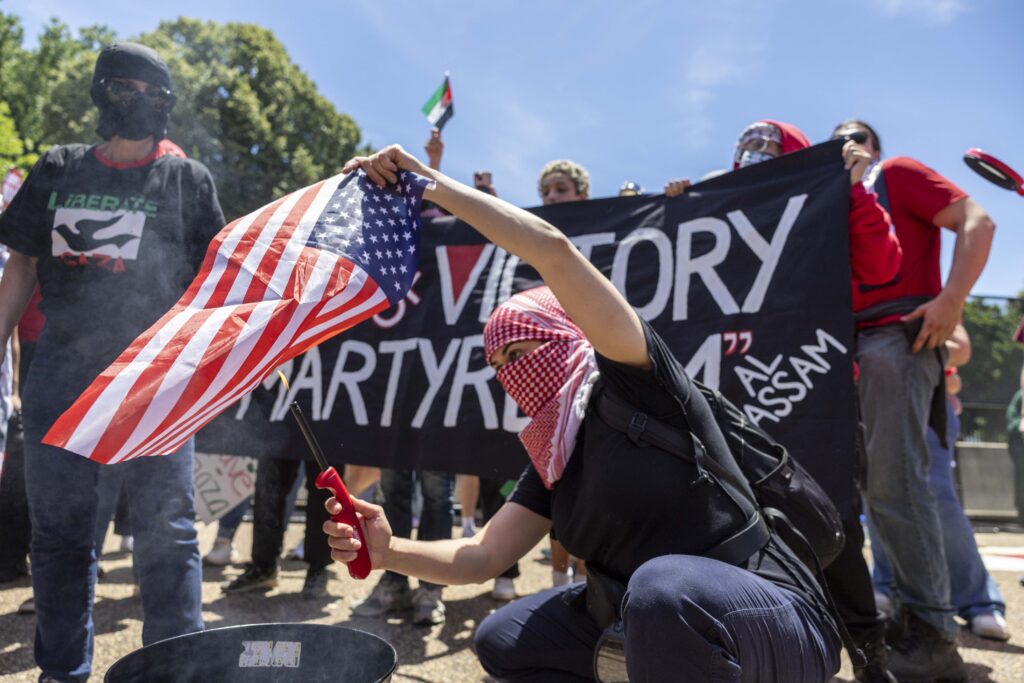 A man wearing a head and face covering crouches as he lights an American flag on fire over a bucket.