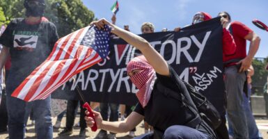 A man wearing a head and face covering crouches as he lights an American flag on fire over a bucket.