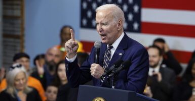 President Joe Biden in a blue suit raises a finger while speaking in a microphone