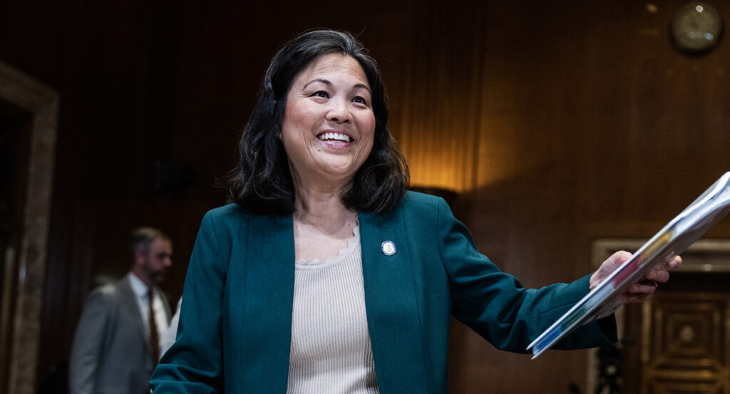 Acting Labor Secretary Julie Su smiles while holding out a binder and wearing a gray sport coat over a blouse