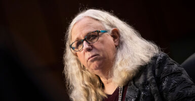 Rachel Levine in a dark blouse with masculine features, long blond hair, and glasses