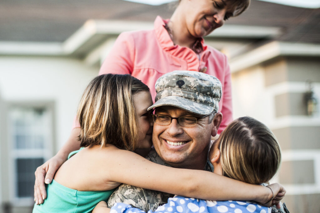 Two children hug their father who is wearing an army uniform while his wife stands over them.