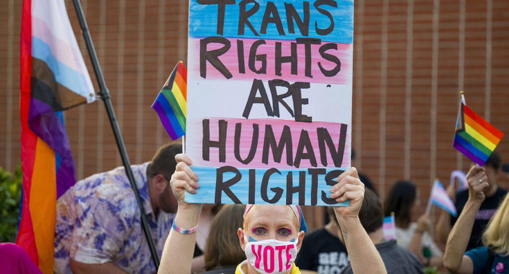 Woman holds sign reading "Trans Rights Are Human Rights" while wearing a face mask with the text "Vote."