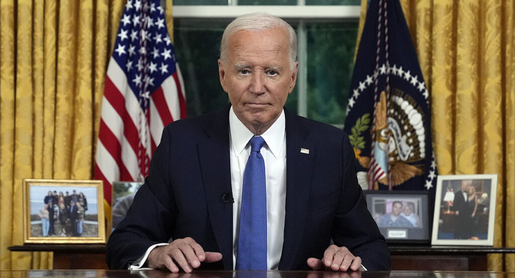 President Joe Biden in a black suit with a blue tie in the Oval Office