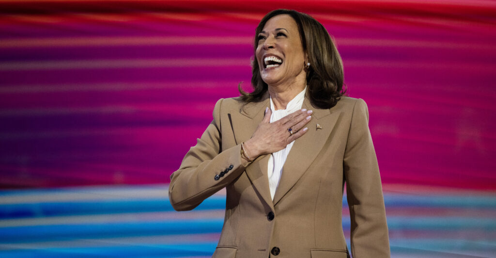 Kamala Harris on stage in a light brown suit smiling with her hand over her heart