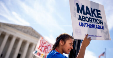 A pro-life supporter stands on a lamppost and holds a sign that reads “MAKE ABORTION UNTHINKABLE” in front of the Supreme Court.