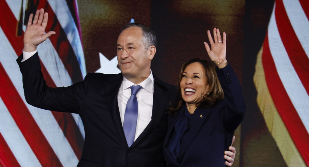 Doug Emhoff in a black suit hugs Kamala Harris in a black suit, as both stand in front of American flags.