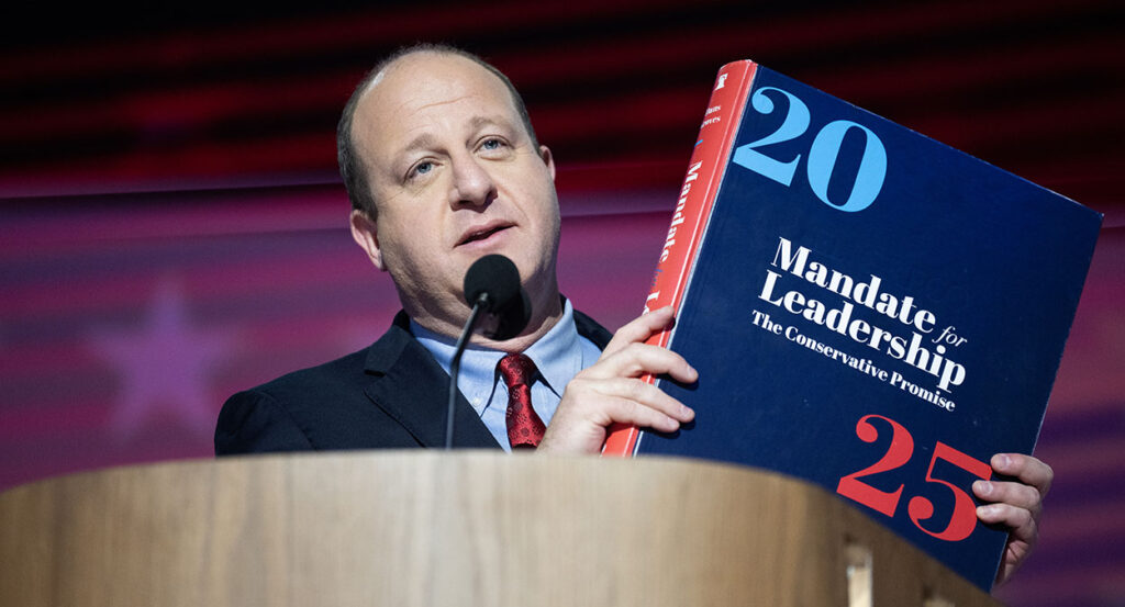 Jared Polis, in a blue suit, holds the Mandate for Leadership Project 2025 book