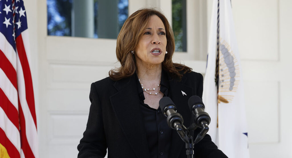 Kamala Harris in a black suit stands behind a microphone in front of an American flag.