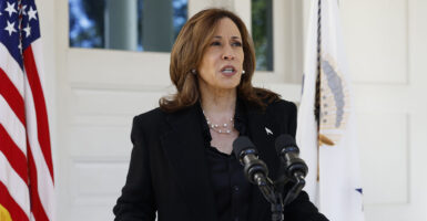 Kamala Harris in a black suit stands behind a microphone in front of an American flag.
