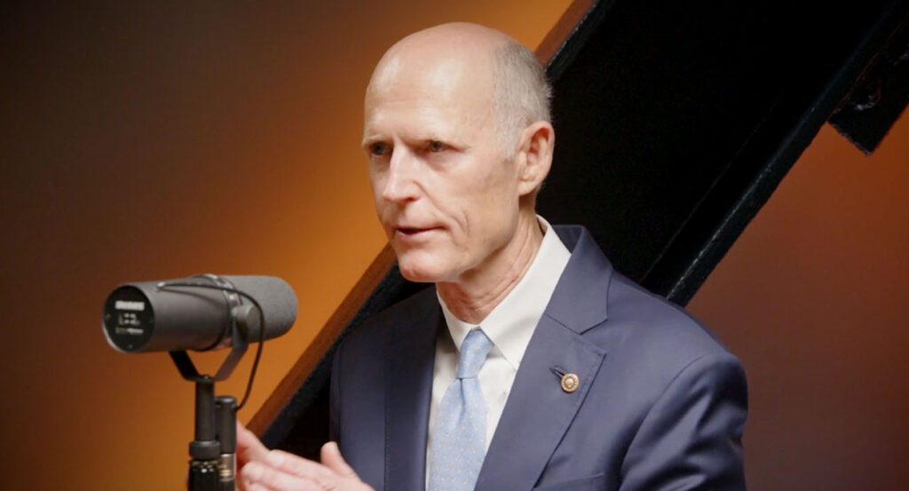 Rick Scott in a grey suit speaks in front of a microphone