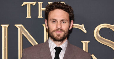 Charlie Vickers stands in a suit in front of the words "The Rings"