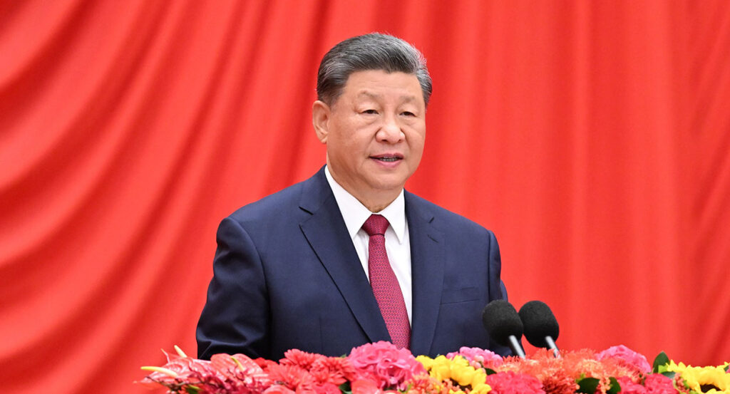 Chinese Premier Xi Jinping in a blue suit speaks in front of a red background with flowers in front of him