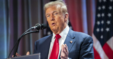Donald Trump in a blue suit and red tie with a flag in the background speaks at a microphone