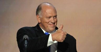 Tom Homan on stage at a lectern smiling and giving a thumbs-up to the audience.