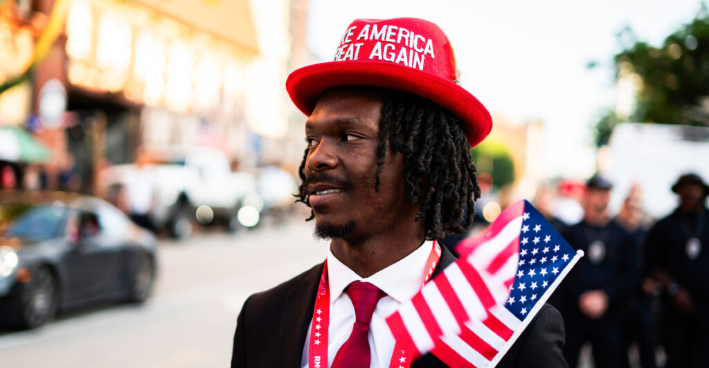 A black Trump supporter stands outside the Republican National Convention on July 17 wearing a red "Make America Great Again" hat and waving an American flag.