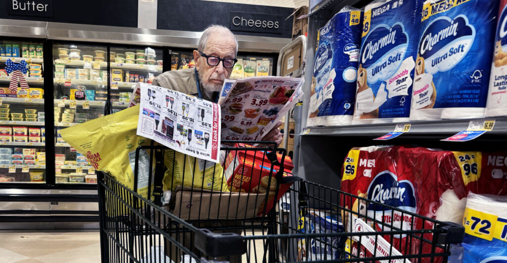 An older man wearing dark-rimmed glasses looks at a coupon book as he pushes a black grocery cart.