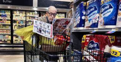 An older man wearing dark-rimmed glasses looks at a coupon book as he pushes a black grocery cart.