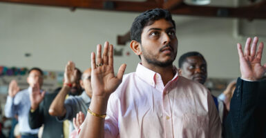 A young man with black hair and brown skin wears a light pink shirt as he raises his right hand with a gold ring on it during his naturalization ceremony.