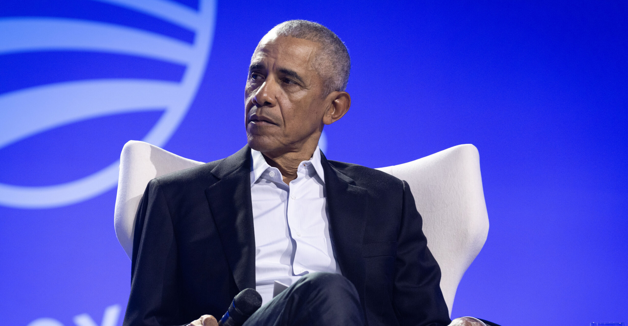 Barack Obama, seated, wears a dark blue suit and white dress shirt as he holds a microphone in his right hand.