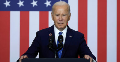 President Joe Biden squints as he stands behind a podium in a navy blue suit, the American flag in the background.