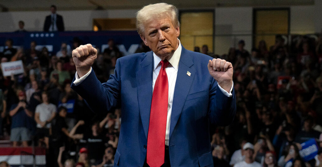 Donald Trump, in a navy suit and red tie, dances with his fists in the air on stage.